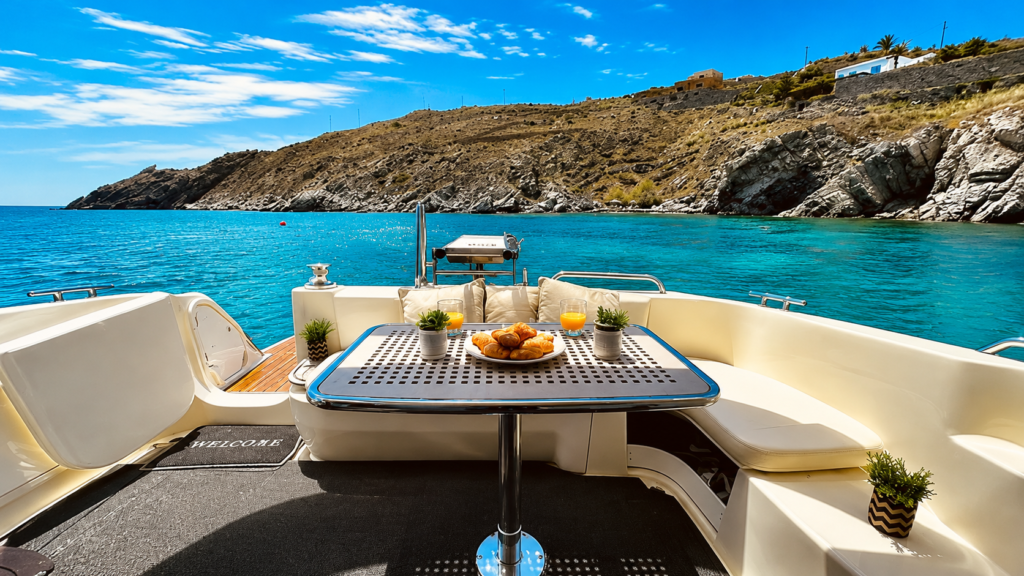 Luxury boat interior with a breakfast table, pastries, and drinks overlooking turquoise water and a rugged coastal landscape.