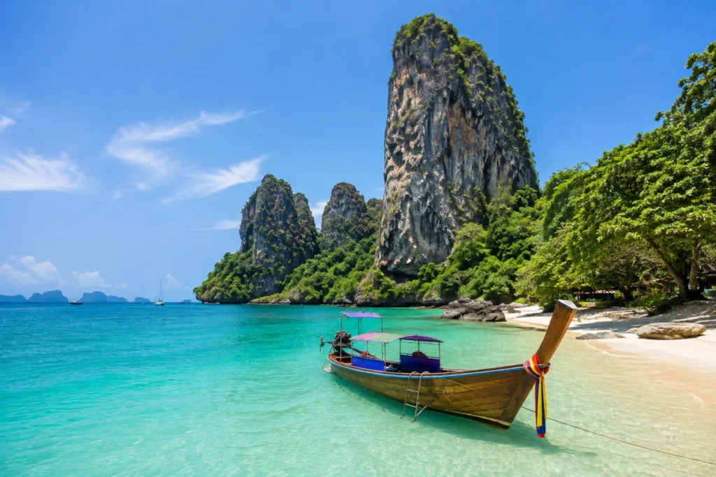 Long-tail boat anchored in crystal-clear turquoise water beside a tropical beach and towering limestone cliffs under a bright blue sky.